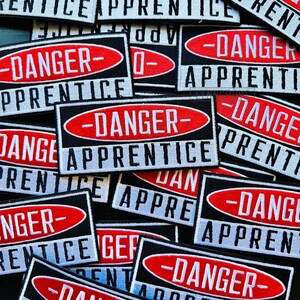 Puede incluir: Un mont&oacute;n de parches negros y rojos con el texto "Danger - Apprentice" en blanco. Los parches est&aacute;n hechos de tela y tienen un borde cosido.