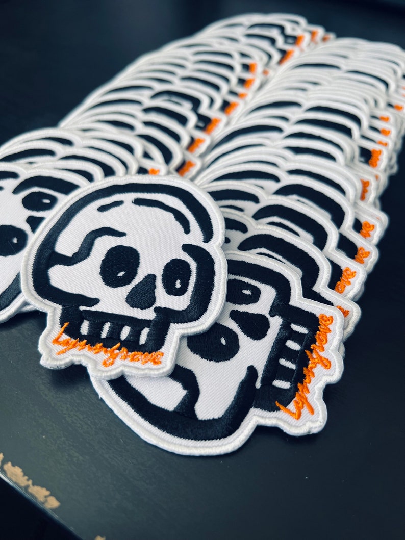 May include: A stack of white fabric patches with black embroidered skull designs and the text "Lazy Bones" in orange thread.
