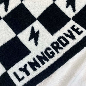 K&ouml;nnte beinhalten: Schwarze und wei&szlig;e karierte Fleece-Decke mit dem Wort "LYMNGROVE" in wei&szlig;en Buchstaben.