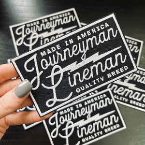 May include: Close-up of four black fabric patches with white embroidered text. The text reads "Made in America Journeyman Lineman Quality Breed".