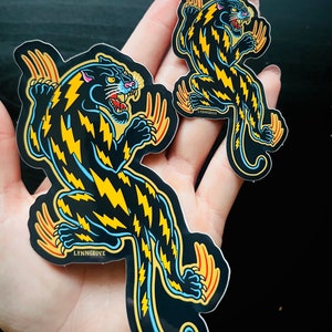 May include: Two black panther stickers with yellow lightning bolt patterns. The larger sticker is 5 inches tall and the smaller sticker is 3 inches tall. Both stickers have the text "LYNNGROVE" printed on them.