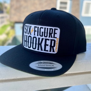 Könnte beinhalten: Schwarze Baseballkappe mit einem schwarzen und weißen gestickten Aufnäher. Der Aufnäher lautet "Six Figure Hooker" mit einem Blitzsymbol. Die Kappe ist eine Snapback der Marke "The Authentic".