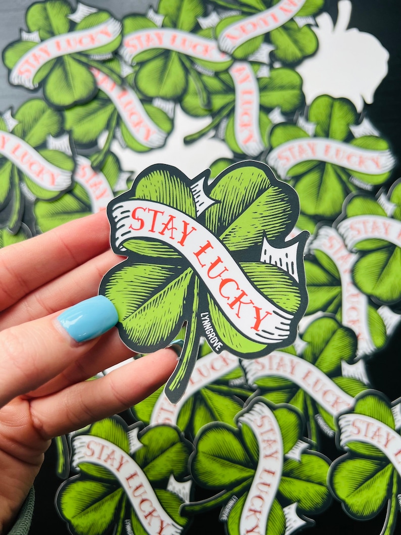 Pu&ograve; includere: Un adesivo verde a forma di trifoglio a quattro foglie con uno striscione bianco che dice "Stay Lucky" in lettere rosse. L'adesivo &egrave; in una mano e ci sono molti altri adesivi simili sullo sfondo.