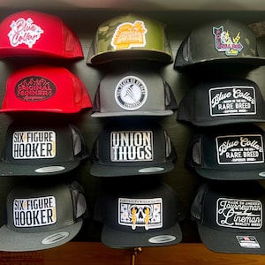 Puede incluir: Una colección de 10 gorras de béisbol negras, grises y rojas con varios parches y logotipos bordados. Los parches presentan frases como "Blue Collar", "Six Figure Hooker", "Union Thugs", "Original Sinners" y "Till Death Do Us Part".