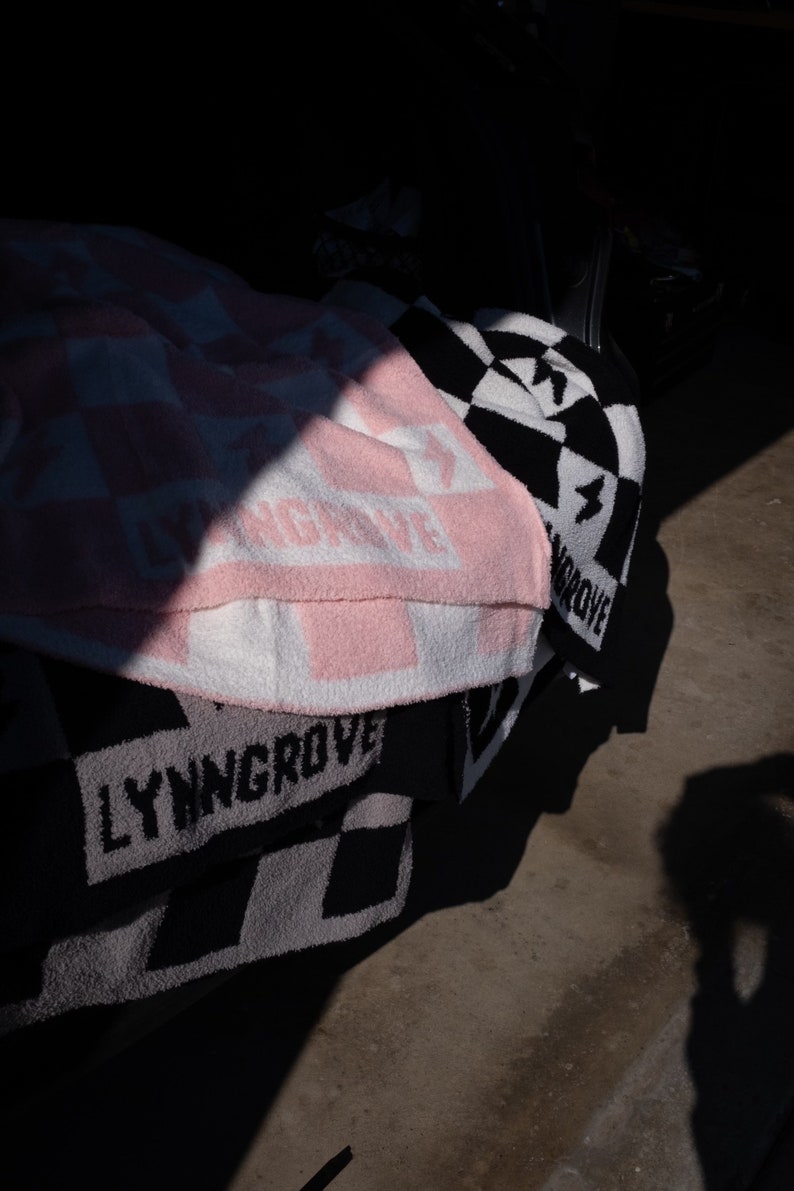 K&ouml;nnte beinhalten: Ein Stapel rosa, wei&szlig; und schwarz karierter Decken mit dem aufgedruckten Text "LYNNGROVE".