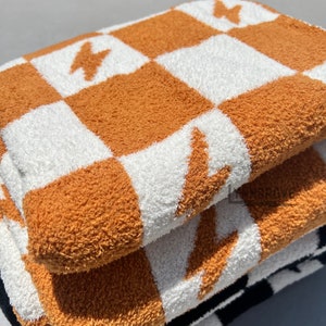 K&ouml;nnte beinhalten: Ein Stapel von drei Pl&uuml;schhandt&uuml;chern mit einem karierten Muster in Orange und Wei&szlig;. Die Handt&uuml;cher haben ein Blitz-Design.