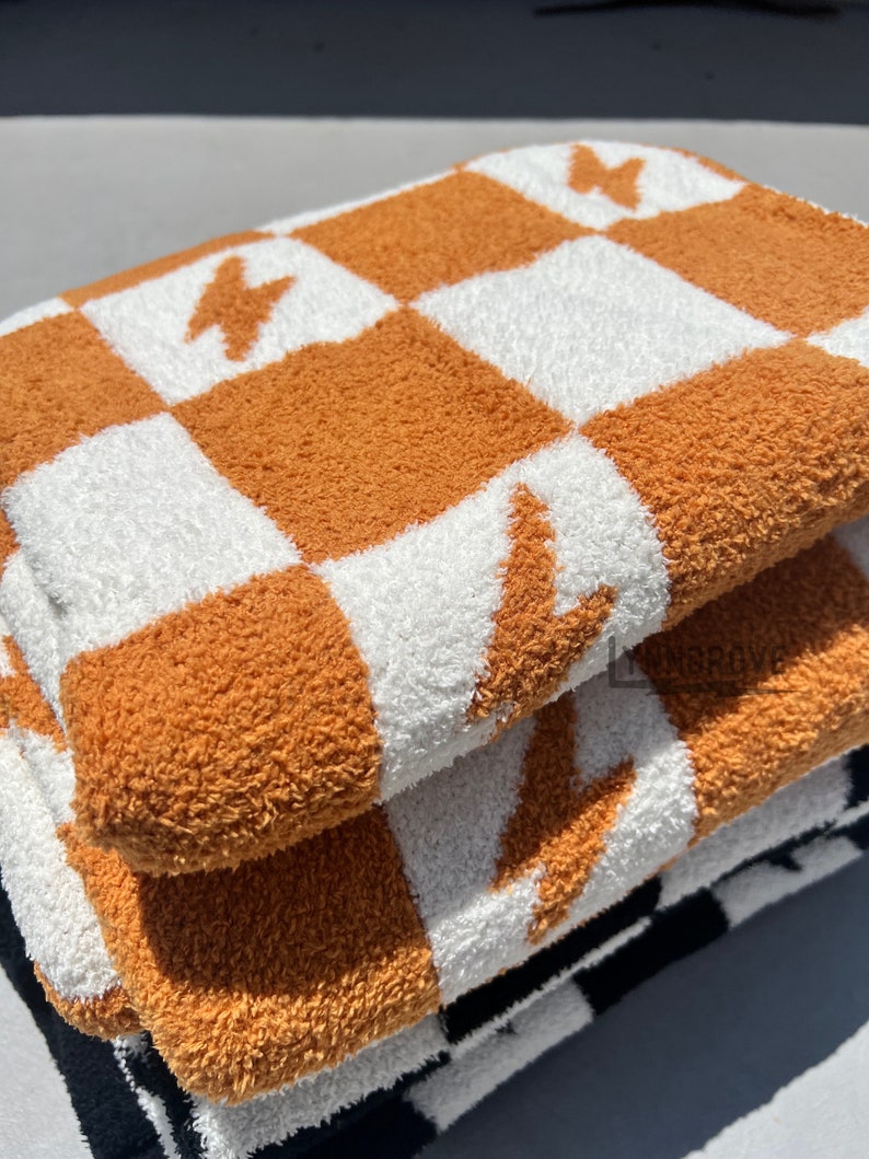 K&ouml;nnte beinhalten: Ein Stapel von drei Pl&uuml;schhandt&uuml;chern mit einem karierten Muster in Orange und Wei&szlig;. Die Handt&uuml;cher haben ein Blitz-Design.