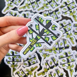 Può includere: Un primo piano di una toppa bianca con una silhouette nera di un albero con banconote verdi appese ai rami. Il testo "MONEY TREE" è scritto in bianco su sfondo nero sulla toppa.