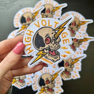 Puede incluir: Parche bordado con un diseño de calavera, un rayo y las palabras "HIGH VOLTAGE". La calavera es de color beige con un parche negro en la cabeza y ojos rojos. El rayo es amarillo. El texto "LYNNGROVE" también está incluido.