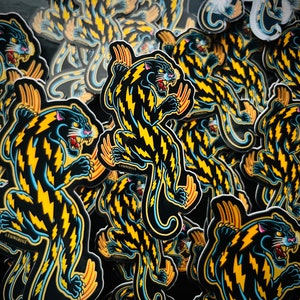 May include: A stack of black and yellow panther stickers with lightning bolt patterns. The stickers have a blue outline and the text "LYINGROVE" is visible on some of the stickers.