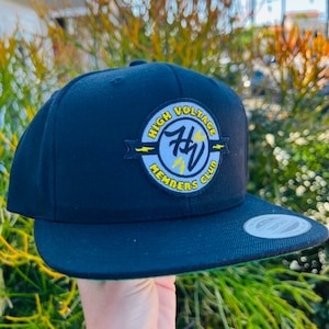 May include: A black snapback hat with a flat brim. The hat features a circular patch with the words "HIGH VOLTAGE MEMBERS CLUB" in yellow and white, with a lightning bolt design. The hat is held up in front of a green and yellow leafy background.