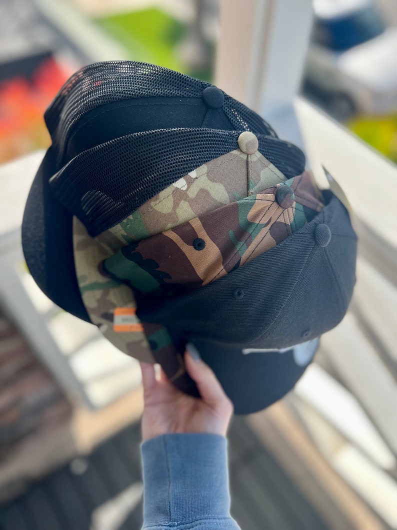 May include: A stack of four baseball caps. The top cap is black with a mesh back. The second cap is black with a camouflage pattern. The third cap is black with a mesh back. The bottom cap is black with a white logo.