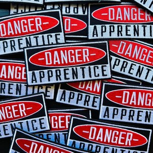 Puede incluir: Un mont&oacute;n de parches de tela negros y blancos con texto rojo y blanco que dice "Peligro - Aprendiz".