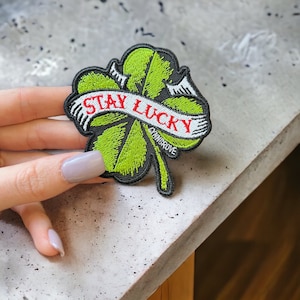 May include: Green four-leaf clover patch with a white banner that reads "Stay Lucky" in red lettering. The patch is embroidered with black stitching and has a small "Cymnsrove" label.