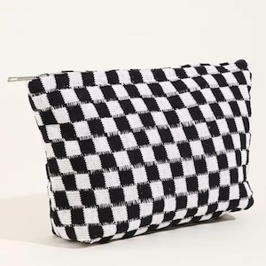 May include: A black and white checkered pattern pouch with a silver zipper. The pouch is rectangular and made of a woven fabric. The black and white squares are evenly sized and create a bold, graphic design. The zipper runs along the top edge.