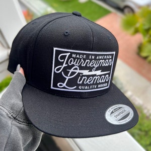 May include: Black and white Journeyman Lineman snapback hat with a white patch that says "Made in America Journeyman Lineman Quality Breed".