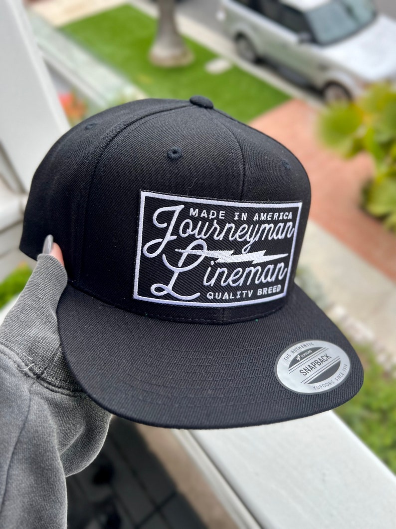 May include: Black and white Journeyman Lineman snapback hat with a white patch that says "Made in America Journeyman Lineman Quality Breed".