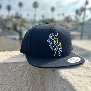 May include: Black fitted baseball cap with a yellow lightning bolt and a white outline of a woman sitting on the bolt. The text "Lightning" is embroidered on the cap.
