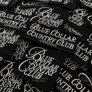 May include: A close-up of several black fabric patches with white embroidered text that reads "Blue Collar Country Club" and "Lynn Grove".