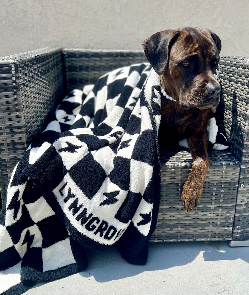 K&ouml;nnte beinhalten: Ein brauner und schwarzer Hund liegt auf einer schwarz-wei&szlig;en karierten Decke mit dem Text "LYNGROW" auf der Decke. Der Hund liegt auf einem Korbstuhl.