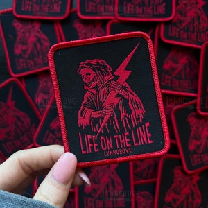 Op de afbeelding: Rode en zwarte geborduurde patch met een skelet dat een bliksem vasthoudt. De patch heeft de tekst "Life on the Line" en "Lynngrove".