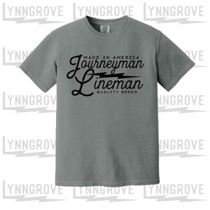 May include: A grey short-sleeve t-shirt featuring black text that reads MADE IN AMERICA JOURNEYMAN LINEMAN QUALITY BREED with a lightning bolt graphic between the words Journeyman and Lineman, printed in a mix of block and script fonts.