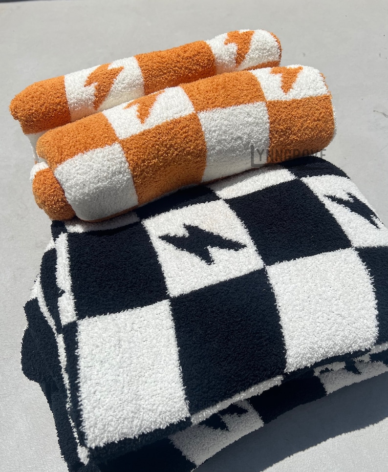 K&ouml;nnte beinhalten: Zwei orange-wei&szlig;e karierte Decken mit einem Blitz-Design, &uuml;bereinander gestapelt auf einer schwarz-wei&szlig;en karierte Decke mit einem Blitz-Design.