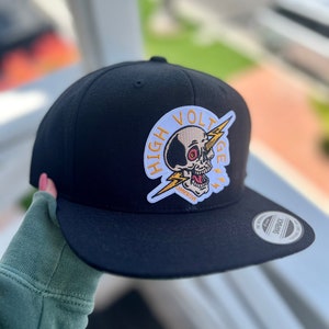 Può includere: Cappellino da baseball nero con una toppa circolare bianca cucita sul davanti. La toppa rappresenta un teschio bianco e nero con un fulmine e il testo "HIGH VOLTAGE" in giallo.