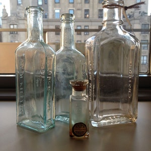May include: Four glass bottles, three are tall and clear, one is short and clear with a cork stopper. The tall bottles have labels with text that is difficult to read. The bottles are on a light brown surface.