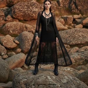 May include: A woman in a black dress and sheer cape stands on a large rock. The dress has a lace skirt and is paired with black boots and layered necklaces. The background features large, weathered rocks.