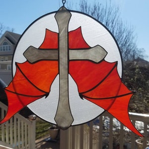 May include: A stained glass window hanging with a silver cross and red wings. The cross is in the center of the circle and the wings are on either side of it.