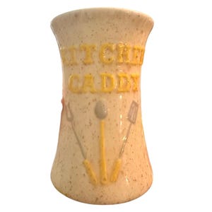 May include: Cream-colored kitchen caddy with a speckled finish. The words "KITCHEN CADDY" are in raised yellow letters. Utensil outlines are also featured. A spoon, spatula, and fork are depicted in yellow and gray.