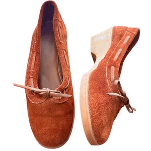 May include: Pair of rust-colored suede shoes with a low heel. The shoes feature a rounded toe, decorative stitching, and a tie detail at the vamp. The heel is approximately 2 inches high.