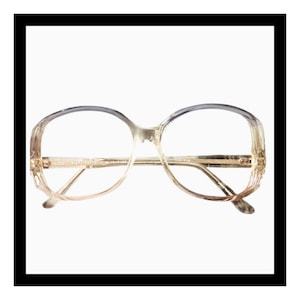 May include: Vintage oversized eyeglasses with a clear, light brown frame and gold-toned metal accents. The lenses are clear, and the top of the frame has a gradient from light brown to a darker gray-blue. The glasses are set against a white background.