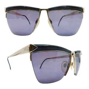 May include: A pair of black and gold sunglasses with a geometric design. The sunglasses have a dark grey lens and a gold metal frame.