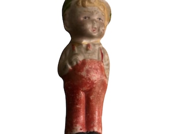 May include: A vintage porcelain doll with a green hat, blonde hair, and red overalls. The doll has a painted face with blue eyes and a light brown complexion.