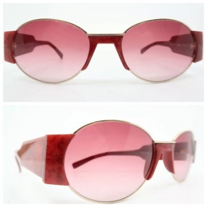 May include: Oval-shaped sunglasses with a gradient pink lens and a gold-colored frame. The temples and the upper frame are a dark red color. The sunglasses have a retro, vintage style.