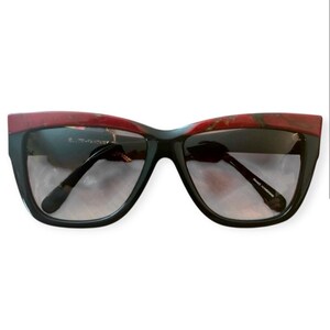 May include: Black cat-eye sunglasses with a red and brown marbled top frame. The lenses have a gradient tint, dark at the top and fading to lighter at the bottom. The text "E...-FANTASY" is visible on the left lens. The sunglasses are a stylish accessory.