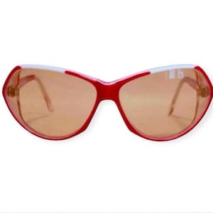Op de afbeelding: Rode zonnebril met een witte accent op de bovenrand. De glazen zijn lichtbruin van kleur. De zonnebril heeft een ongewone, hoekige vorm en dunne, goudkleurige pootjes.