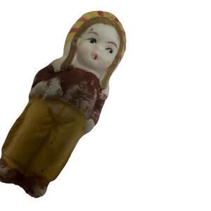 Puede incluir: Una peque&ntilde;a figurita de porcelana de una ni&ntilde;a que lleva un sombrero amarillo y rojo y un vestido marr&oacute;n y amarillo.