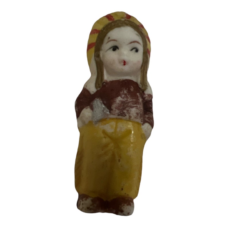 Puede incluir: Una peque&ntilde;a figurita de porcelana de una ni&ntilde;a que lleva un sombrero amarillo y rojo, una camisa marr&oacute;n y pantalones amarillos.