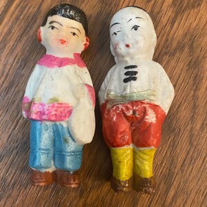 May include: Two porcelain dolls with painted faces and clothing. The doll on the left is wearing a white shirt with pink and yellow accents, blue pants, and black shoes. The doll on the right is wearing a white shirt with black stripes, red pants, yellow shoes, and a green sash.