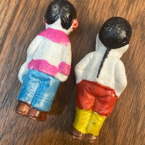 May include: Two vintage porcelain dolls with painted details. One doll is wearing a pink and white striped shirt, blue pants, and brown shoes. The other doll is wearing a white shirt, red pants, yellow pants, and brown shoes.