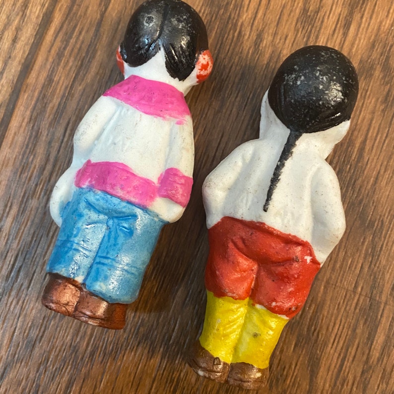 May include: Two vintage porcelain dolls with painted details. One doll is wearing a pink and white striped shirt, blue pants, and brown shoes. The other doll is wearing a white shirt, red pants, yellow pants, and brown shoes.