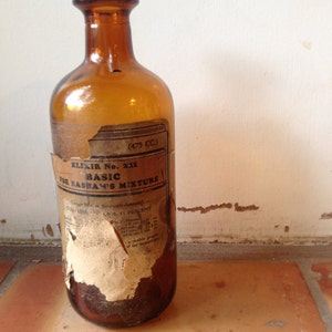 May include: An amber-colored glass bottle with a label that reads "ELIXIR No. 211 BASIC FOR BASHAM'S MIXTURE." The label is partially torn and aged. The bottle has a capacity of 475 cc.