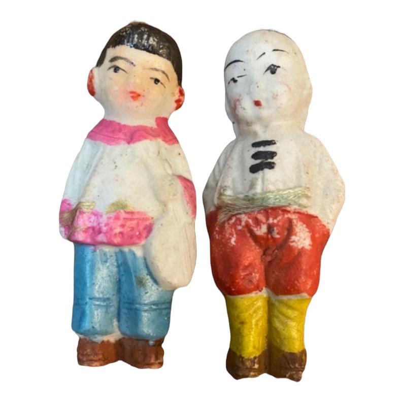 May include: Two small porcelain figurines of people wearing traditional clothing. The figurine on the left is wearing a white shirt with pink accents, blue pants, and a white hat. The figurine on the right is wearing a white shirt with black accents, red pants, and a yellow hat.