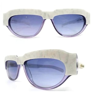 May include: Stylish sunglasses with a marbled white frame and purple lenses. The lenses have a gradient effect, transitioning from a darker shade at the top to a lighter shade at the bottom. The frames have a unique, angular design.