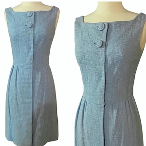 May include: A pale blue sleeveless dress with a square neckline and two matching buttons. The dress features a fitted bodice and a pleated skirt, made from a textured fabric. The dress is displayed on a mannequin.
