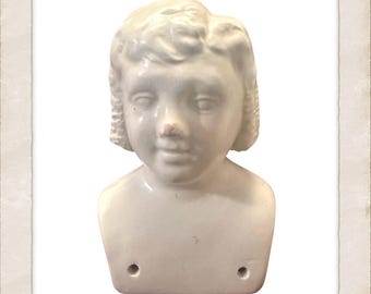 Op de afbeelding: Een vintage biscuit poppenhoofd, met een serene uitdrukking en kort, krullend haar. Het porseleinen hoofd is gebroken wit met subtiele schaduwen en een kleine vlek op de neus. Twee gaten zijn zichtbaar aan de basis van de nek. Witte achtergrond.