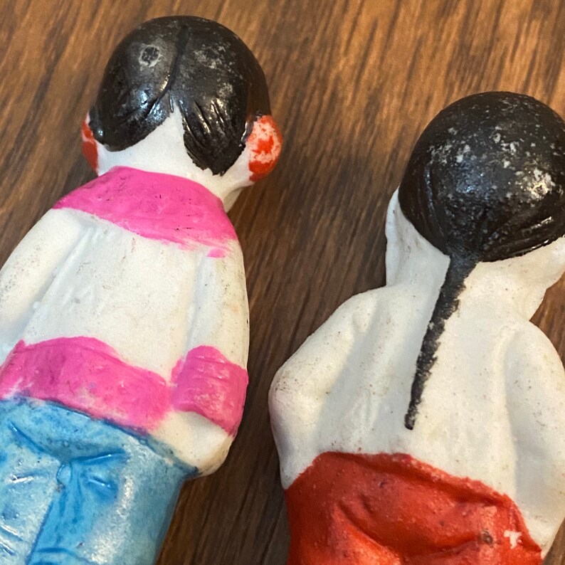 May include: Two porcelain dolls with black hair and painted features. One doll is wearing a pink and white striped shirt with blue pants. The other doll is wearing a red skirt.